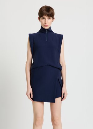 Marineblaues ärmelloses Oberteil mit hohem Kragen und Reißverschlussdetail, kombiniert mit einem passenden Wickelrock mit seitlichem Schlitz.