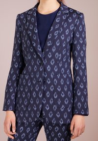 Mörkblå kavaj med blåmönstrad design, strukturerad form, slagkrage, två framfickor och enskild knappstängning.