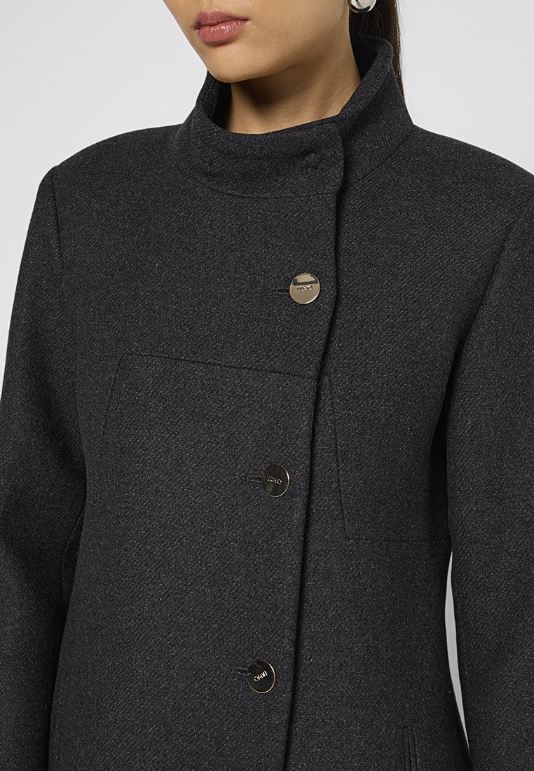 Donna che indossa un cappotto di lana grigio scuro con collo alzato e tre bottoni metallici lucidi visibili sul davanti.