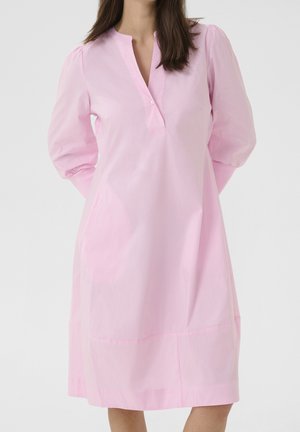 Femme portant une robe ample rose clair longueur genoux avec des manches longues bouffantes et un col en V avec un seul bouton, debout devant un fond uni.