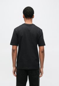 Melna kokvilnas T-krekls ar īsām piedurknēm un apaļu kaklu, mīksta tekstūra, vienkāršs dizains, valkāts kopā ar melnām džinsu biksēm, redzams no mugurpuses.