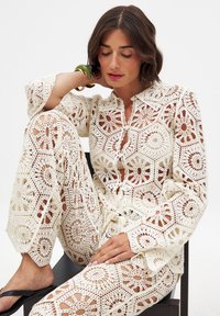 Completo all'uncinetto bianco composto da una camicia con colletto e chiusure a bottone e pantaloni a gamba larga, che mostrano motivi floreali e una texture a trama aperta.