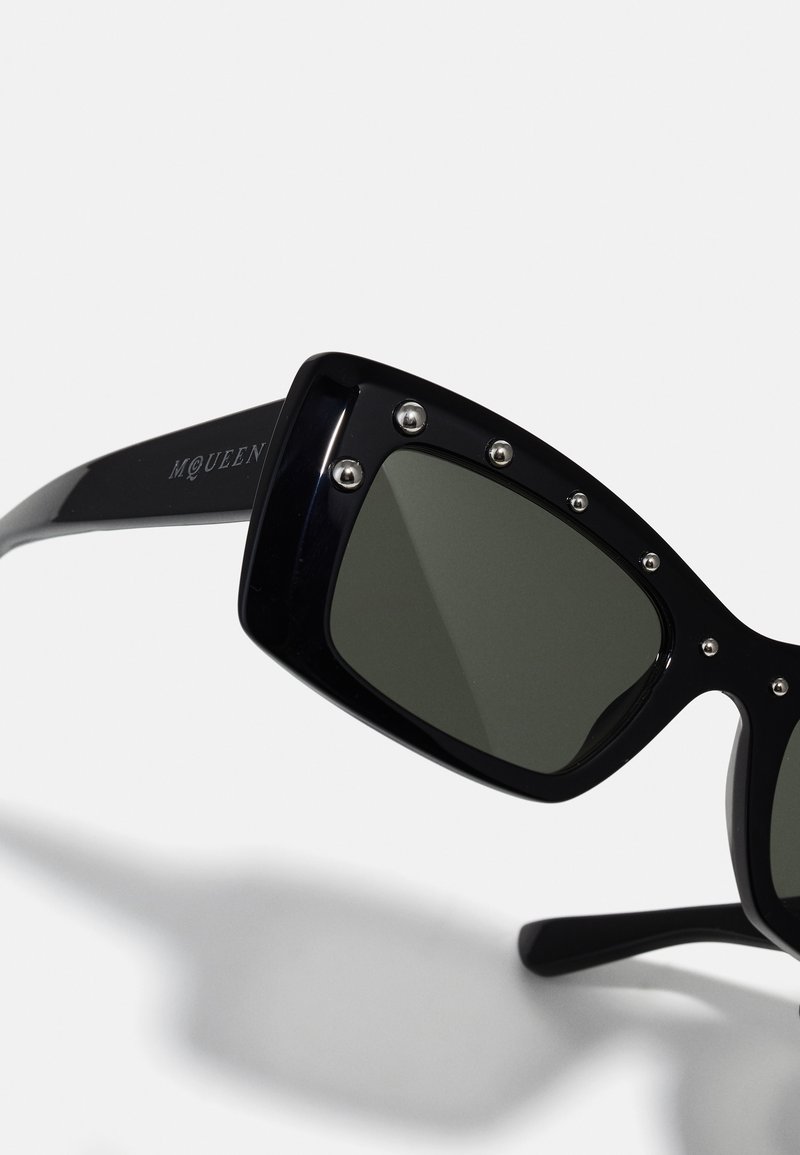 Occhiali da sole rettangolari neri con lenti scure, borchie di metallo sul telaio e il testo "McQueen" sulla tempia. Finitura liscia e lucida.