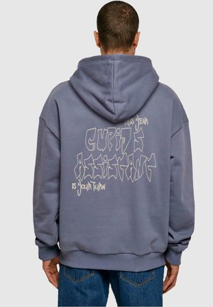 Mann trägt einen blau-grauen Hoodie mit Graffiti-Stil Text „Dieses Jahr ist Amor Assistent, du bist dran“ auf dem Rücken, kombiniert mit blauen Jeans.