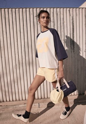 Femme marchant à l'extérieur portant un t-shirt blanc et bleu marine avec le numéro 23, un short jaune, des chaussettes noires, des chaussures blanches, et portant un sac bleu marine et une casquette jaune.