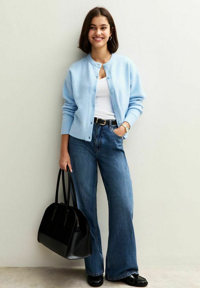 Jeune femme portant un cardigan bleu clair, un top blanc, un jeans large, des chaussures noires, tenant un grand sac à main noir, souriant devant un mur uni.