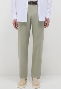 Mann in beigen Hosen, einem weißen offenen Hemd, einem braunen geflochtenen Gürtel und beigen Slippers, stehend vor einem schlichten weißen Hintergrund.