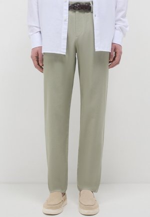 Uomo che indossa pantaloni beige, una camicia bianca aperta, una cintura intrecciata marrone e scarpe beige slip-on, in piedi contro uno sfondo bianco semplice.