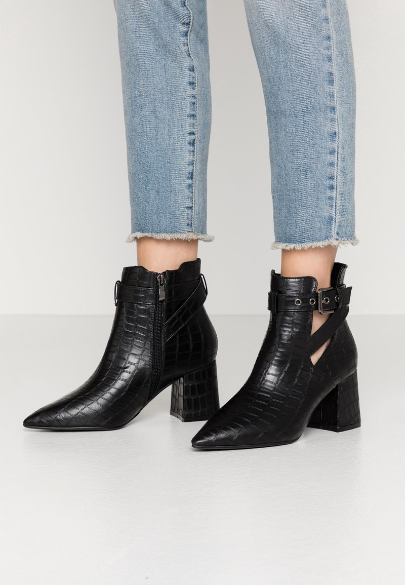 Bottines noires à effet croco, talons carrés, bouts pointus, fermeture éclair latérale, brides à boucle, portées avec un jean bleu clair coupé et effiloché.