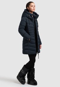 Temno modra oblazinjena jakna z visokim ovratnikom in nastavljivimi vrvicami, opremljena z žepki z zadrgami in ravnim krojem. Nošena s črnimi škornji.