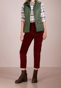 Grüne gesteppte Weste über einem karierten Hemd, kombiniert mit burgunderroten Cordhosen und braunen Schnürstiefeln. Strukturierte Stoffe und kontrastreiche Farben.