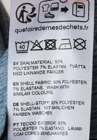 Zorglabel met een witte achtergrond die wasinstructies en de samenstelling van de stof weergeeft: 93% polyester, 7% elastaan, in meerdere talen.