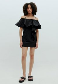 Schwarzes schulterfreies Kleid mit Rüschendekolleté und asymmetrischem Vorderteil. Glattes Material, figurbetonte Silhouette und kombiniert mit schwarzen Sandalen.