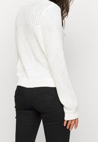 Vit stickad tröja med ett texturerat mönster, ribbade muddar och fåll. Buren över svarta tajta jeans, med en avslappnad bakifrån vy.