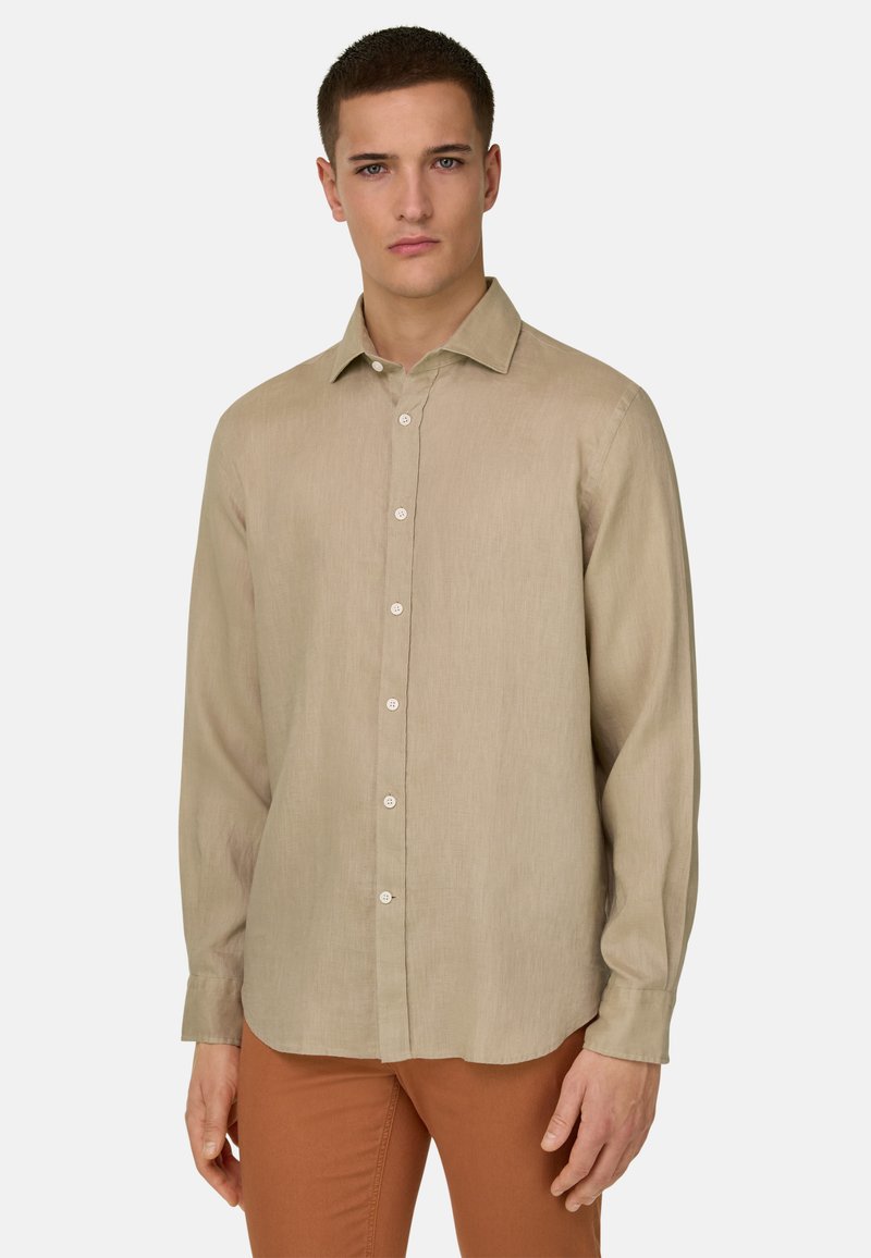 Jeune homme portant une chemise beige à manches longues boutonnée et un pantalon couleur rouille, debout devant un fond clair uni.