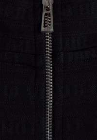 Tissu noir à texture côtelée, avec une fermeture éclair métallique argentée au centre. La fermeture éclair présente des dents visibles et une tirette.