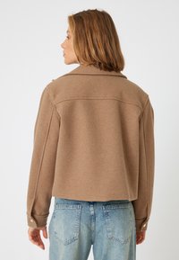 Giacca corta in lana marrone con colletto classico e polsini abbottonati, abbinata a jeans in denim azzurro chiaro. Texture liscia, minimalismo nei dettagli.