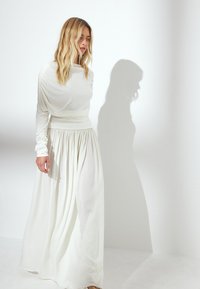 Lungo abito bianco realizzato in tessuto fluente con un corpetto drappeggiato e vita arricciata. Le lunghe maniche e la gonna ampia e plissè creano una silhouette morbida.