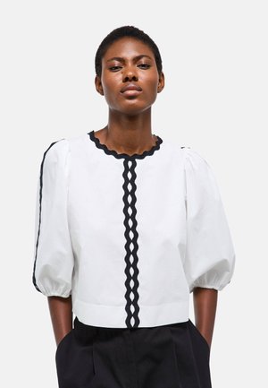 Jeune femme portant un chemisier blanc avec un bord festonné noir et des manches bouffantes, debout les mains dans les poches d'un pantalon noir.