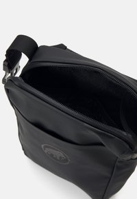Svart crossbody-väska gjord av strukturerad tyg, med dragkedjestängning, justerbar rem och en björnlogotyp på framsidan.