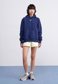 Marineblå hettegenser med en lomme foran, snor, og en hvit Nike-logo. Sammen med lysegule shorts og grå joggesko.
