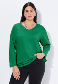 Zielona bluzka z długim rękawem i dekoltem w serek, wykonana z miękkiego materiału, z bocznymi rozcięciami i luźnym krojem. Noszona z ciemnymi jeansami i warstwowymi naszyjnikami.