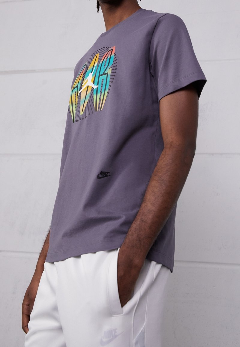 Homme portant un t-shirt Nike violet avec un motif coloré et un pantalon de survêtement Nike blanc, debout une main dans la poche contre un mur gris.