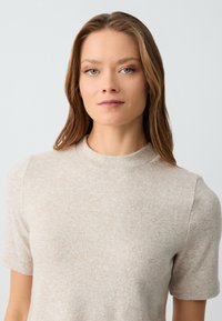 Top corto a maniche corte color beige con scollatura rotonda alta. Il tessuto presenta una leggera texture e varia leggermente di tonalità in tutta la sua superficie.