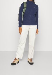 Marineblaue Fleecejacke mit hohem Kragen und Reißverschluss. Kombiniert mit weißen Hosen und beigen Schuhen, die orangefarbene Akzente und schwarze Sohlen haben.