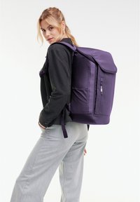 Un sac à dos violet avec un design structuré, des bretelles rembourrées, une poche avant zippée et une texture lisse. Porté sur le dos.