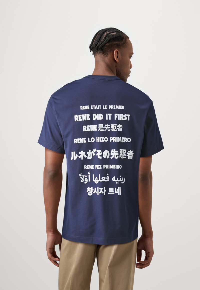 Camiseta de algodón azul marino con grandes letras blancas en varios idiomas en la parte posterior, incluyendo lemas relacionados con "Rene". Mangas cortas, ajuste relajado.