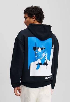 Felpa nera con cappuccio caratterizzata da una grande stampa grafica blu di una figura con occhiali da sole sulla parte posteriore. Tessuto morbido, vestibilità comoda, orlo a coste.