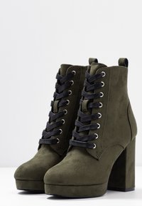 Bottes de cheville en suède synthétique vert olive avec un talon épais, des lacets noirs et des œillets métalliques ; design à bout arrondi et hauteur de cheville.