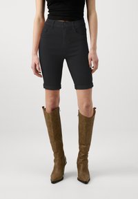 Pantalones cortos de mezclilla negra oscura con puños doblados, combinados con botas de cuero marrón hasta la rodilla con punta afilada y acentos cosidos.