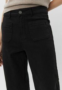 Crne traperice s visokim strukom, prednjim kopčanjem dugmetom i dva stražnja džepa s horizontalnim šavovima. Glatka tekstura tkanine.