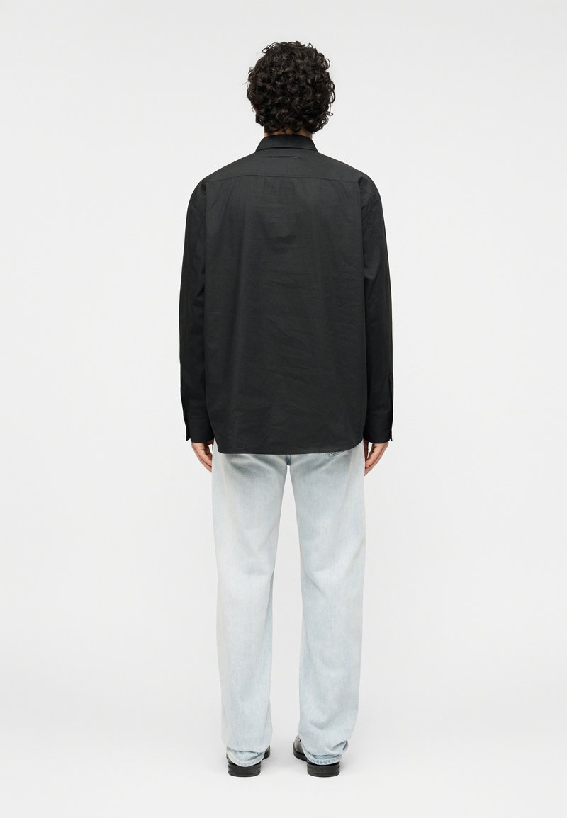 Chemise noire à manches longues avec une coupe droite, portée sur un jean bleu clair à jambes larges et des chaussures noires. Tissu lisse avec peu de détails.