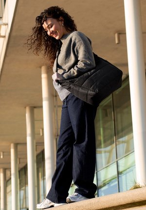 Femme souriante aux cheveux bouclés portant un pull gris, un pantalon foncé, des baskets blanches, et portant un grand sac à bandoulière noir en extérieur.