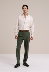 Chemise beige clair à boutons portée par-dessus un sous-vêtement blanc, associée à un pantalon vert foncé et des chaussures noires, présentant une coupe ajustée.