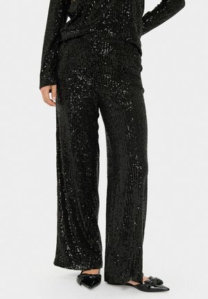 Schwarze, mit Pailletten besetzte, weite Hosen mit strukturiertem Finish. Das Design weist vertikale Reihen von Pailletten und einen glatten Bund auf. Getragen mit schwarzen spitzen Ballerinas.