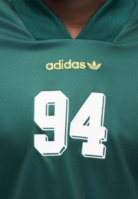 Πράσινη αθλητική φανέλα από πολυεστέρα με λευκό αριθμό "94" και χρυσό λογότυπο "adidas" με τρεις ρίγες πάνω από αυτό. Σχέδιο με κοντό γιακά.