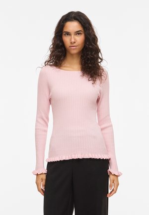 Hosszú, göndör hajú nő világos rózsaszín bordázott pulóverben, fodros mandzsettákkal és szegéllyel, fekete nadrággal, fehér háttér előtt állva.