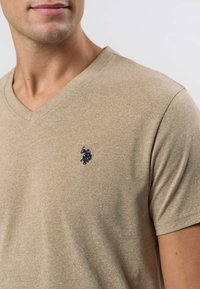 V-Ausschnitt, kurzärmeliges Shirt aus hellbeigem Stoff mit subtiler Textur. Mit kleinem marineblauem Logo auf der linken Brust.