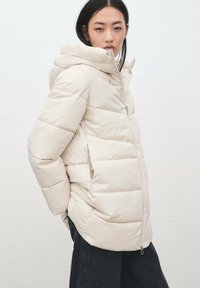 Kurtka puchowa w jasnobeżowym kolorze, z kapturem, poziomym pikowaniem i zamkiem błyskawicznym z przodu. Miękka w dotyku i o luźnym kroju.