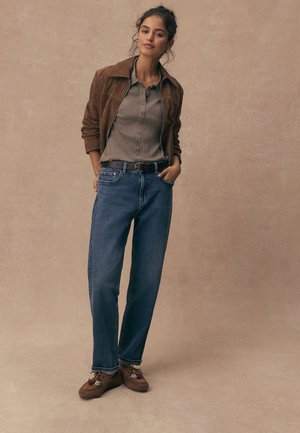 Ung kvinde stående med hænderne i lommerne, iført brun ruskindsjakke, grå skjorte med knapper, blå jeans og brune mokkasiner.