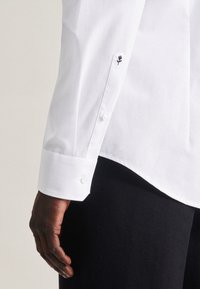 Chemise blanche à manches longues avec poignet à boutons et détail de rose brodé. Texture lisse, coupe ajustée et ourlet arrondi.