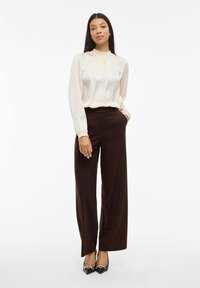 Blusa crema con scollo arricciato e maniche lunghe, abbinata a pantaloni ampi beige a vita alta. Il modello sta in piedi con le mani in tasca.