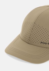 Gorro beige con diseño perforado, visera curva, tela acolchada y costuras en contraste. Presenta un logotipo en el costado.
