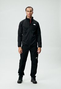 Sort zip-jakke og bukser lavet af slidstærkt stof, med The North Face-logoet på venstre bryst og lår, parret med sorte sko.