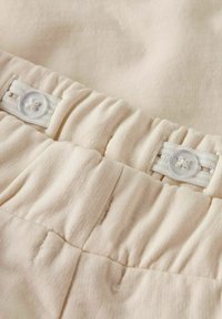 Elastischer beiger Stoffbund mit verstellbaren weißen Knöpfen auf einer weichen Textur.