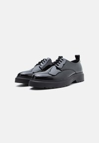 Um par de sapatos derby pretos brilhantes, com atacadores e solas de borracha grossas, contra um fundo branco, angulados para mostrar a frente e os lados.